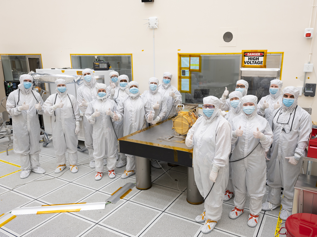 A large group of engineers in full white body coveralls stand surrounding Europa Clipper’s surface dust analyzer. All the engineers are giving a “thumbs up” hand gesture to indicate that the instrument was successfully delivered. Nine engineers stand to the left of a silver workbench, on which the instrument rests, and six stand to the right of the workbench. The instrument is gold colored and about the size of a drum, resting on its side. The instrument is resting upon a silver workbench in the cleanroom. 